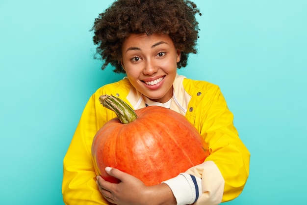 Pleased smiling young Afro American woman embraces big pumpkin, dressed in yellow raincoat, has happy expression, isolated over blue background.