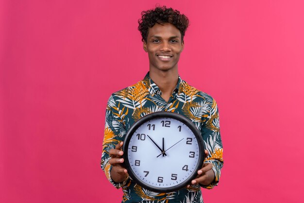 Pleased and smiling young dark-skinned man with curly hair in leaves printed shirt holding wall clock with hands showing time on a pink background