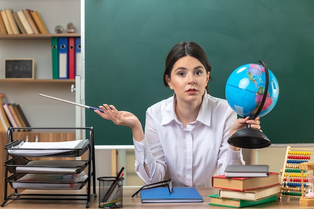 Бесплатное фото Довольно протянув руку молодая учительница, сидящая за столом со школьными инструментами, держащая глобус с указателем в классе
