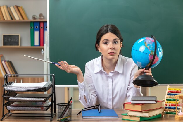 Pleased spreading hand young female teacher sitting at table with school tools holding globe with pointer stick in classroom