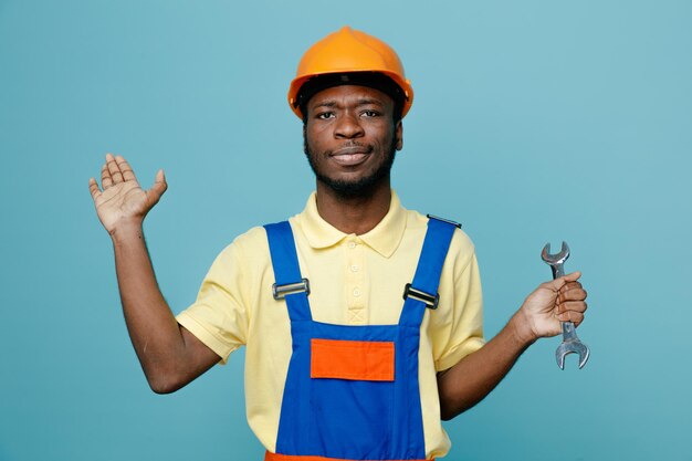 Pleased spreading hands holding open end wrench young african american builder in uniform isolated on blue background