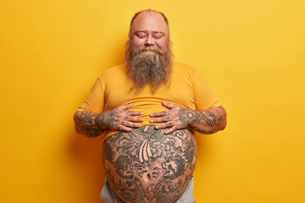 Pleased stout man keeps hands on belly, feels satiety after eating delicious dinner, stands with eyes closed, doesnt care about figure, has hormonal imbalance, isolated on yellow wall.