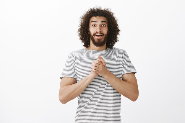 Pleased stunned attractive Eastern guy with beard and stylish curly haircut, holding clasped palms on chest and loosing speech from something positive