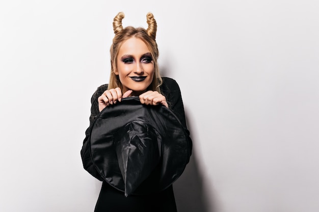 Free photo pleased stylish woman in black outfit celebrating halloween. adorable girl in witch costume preparing for party.