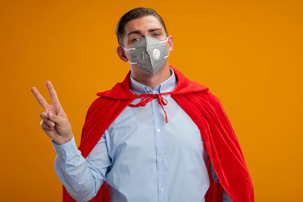 Pleased super hero businessman in protective facial mask and red cape looking at camera showing v-sign standing over orange background