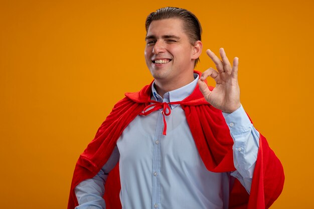 Pleased super hero businessman in red cape looking at camera smiling broadly showing ok sign standing over orange background