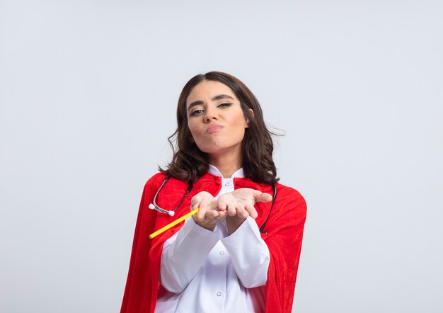 Pleased superwoman in doctor uniform with red cape and stethoscope keeps hands together holding pencil isolated on white wall