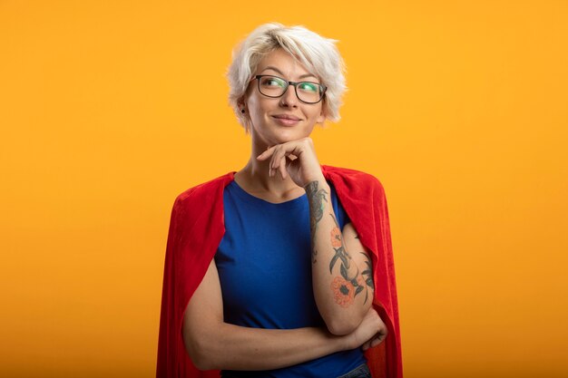 Pleased superwoman with red cape in optical glasses puts hand on chin and looks at side isolated on orange wall