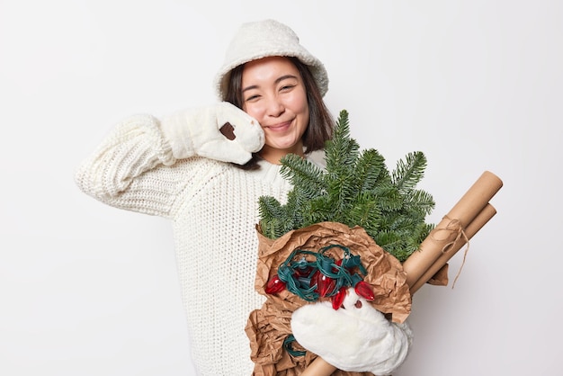 Pleased tender beautiful woman wears warm white knitted sweater mittens and hat for cold winter days holds green spruce branches garland and paper for wrapping presents prepares for new year