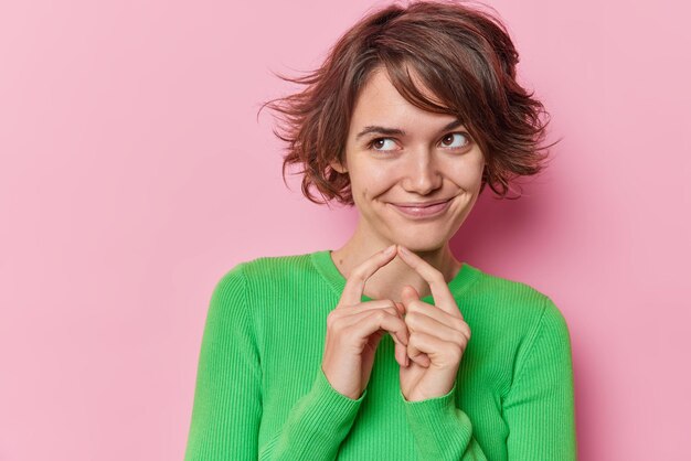 Pleased thoughtful beautiful European woman with short hairstyle keeps index fingers together and schemes something something wears green jumper isolated over pink background has tricky plan