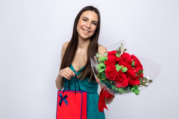 Pleased tilting head beautiful young girl holding bouquet with gift bag