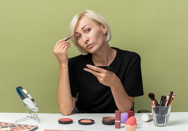 Pleased tilting head young beautiful girl sits at table with makeup tools holding makeup brush isolated on olive green wall