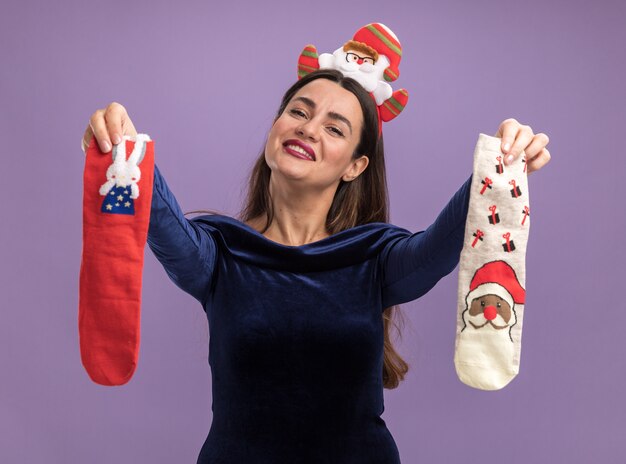 Pleased tilting head young beautiful girl wearing blue dress and christmas hair hoop holding christmas socks isolated on purple background