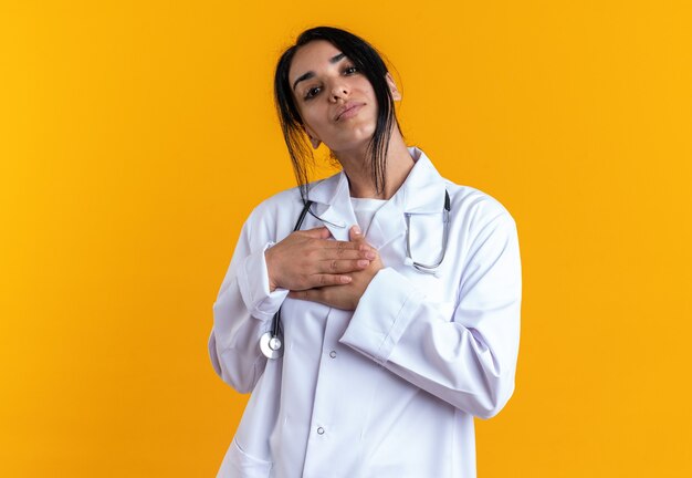 청진 기 노란색 벽에 고립 된 자신에 손을 댔을 의료 가운을 입고 기쁘게 틸팅 머리 젊은 여성 의사
