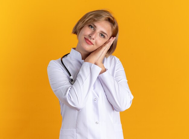 Довольно наклонив голову молодая женщина-врач в медицинском халате со стетоскопом показывает жест сна, изолированный на оранжевой стене