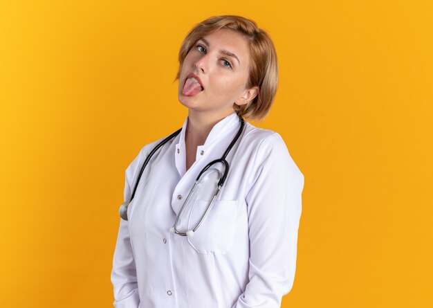 オレンジ色の壁に分離された舌を示す聴診器で医療ローブを身に着けている頭を傾けて喜んで若い女性医師