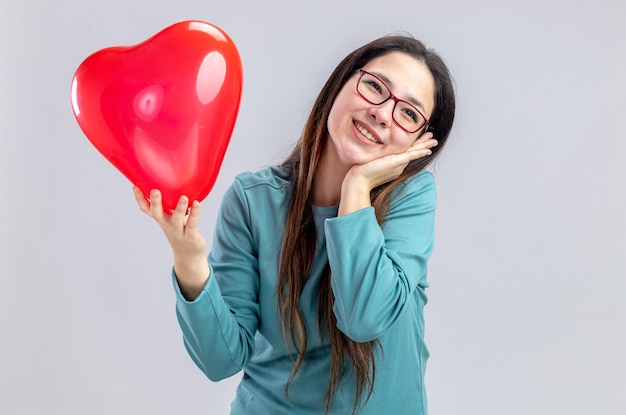 Pleased tilting head young girl on valentines day holding heart balloon putting hand on cheek isolated on white background