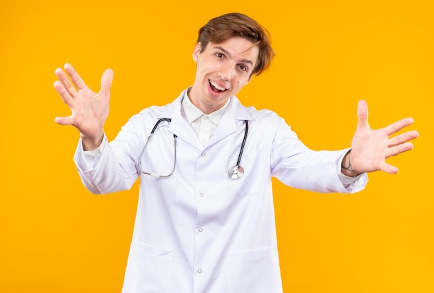 Pleased tilting head young male doctor wearing medical robe with stethoscope holding out hand isolated on orange wall