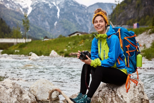 Pleased tourist rests on rocks, holds photocamera for taking shots