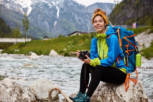 Pleased tourist rests on rocks, holds photocamera for taking shots