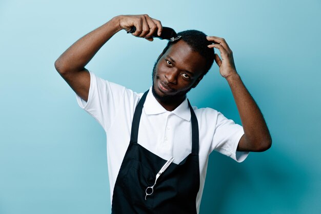 Pleased trimming his hair with hair clippers young african american barber in uniform isolated on blue background