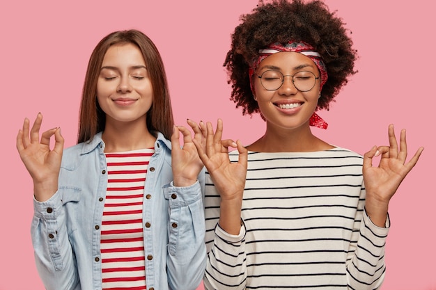 Pleased two women of different race make okay gesture with both hands, close eyes, try to concentrate