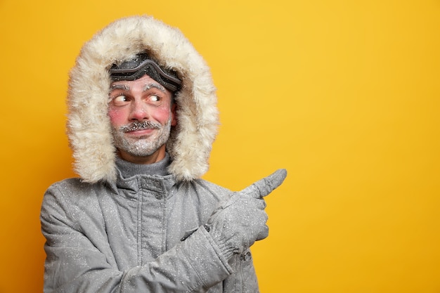 Pleased unshaven European man suggests to participate in ski racing competition wears grey outerwear points at copy space