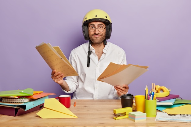 Pleased unshaven guy wears helmet and white shirt, looks through documents at workplace, makes project