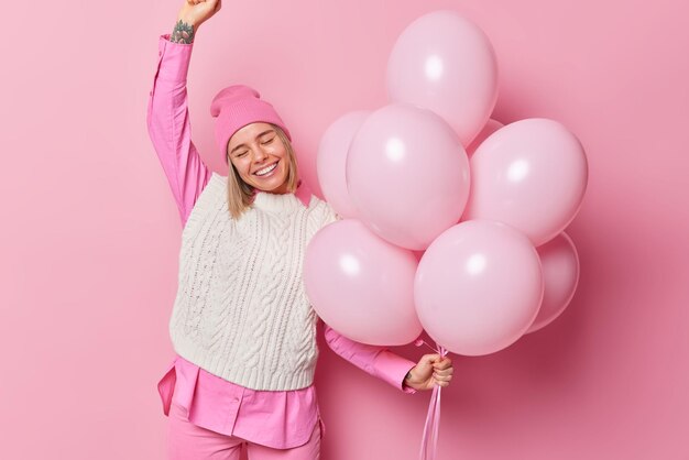 Pleased upbeat woman wears fashionable clothes dances to music on party has festive mood poses against pink background with inflated balloons celebrates birthday with friends Holidays concept