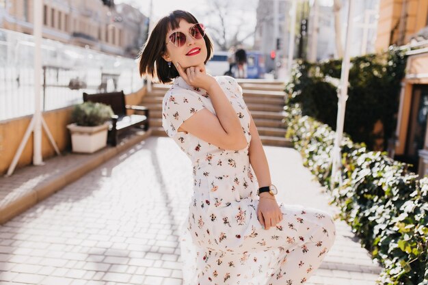 Pleased white woman in trendy summer dress dancing on the street Outdoor photo of smiling female model with short brown hair expressing positive emotions in spring day