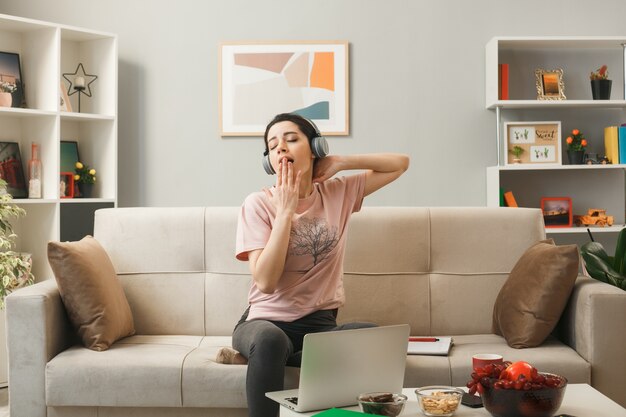 Pleased with closed eyes covered mouth with hand young girl wearing headphones sitting on sofa behind coffee table in living room