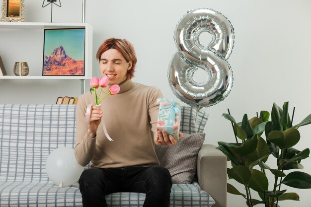 Pleased with closed eyes handsome guy on happy women day holding flowers with present sitting on sofa in living room