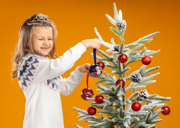 Довольная с закрытыми глазами маленькая девочка стоит рядом с елкой в тиаре с гирляндой на шее и держит елочные шары на оранжевом фоне