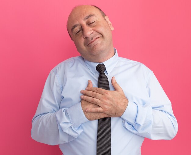 Pleased with closed eyes middle-aged man wearing white t-shirt with tie putting hand on heart isolated on pink wall