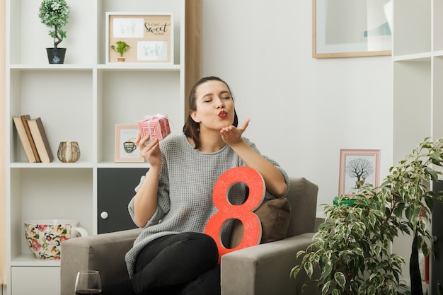 Pleased with closed eyes showing kiss gesture beautiful woman on happy women day holding present sitting on armchair in living room