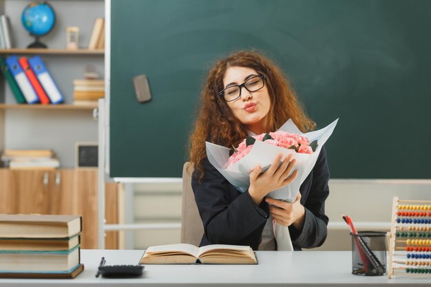 教室で学校の道具を持って机に座っている花の花束を持っている若い女性教師がキスジェスチャーを示す目を閉じて満足