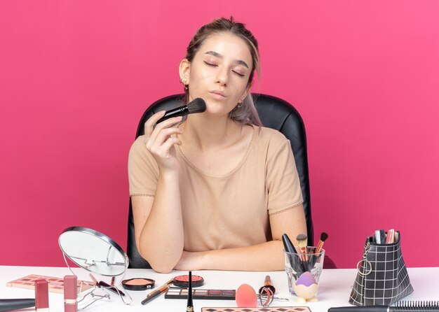 Pleased with closed eyes young beautiful girl sits at table with makeup tools isolated on pink wall