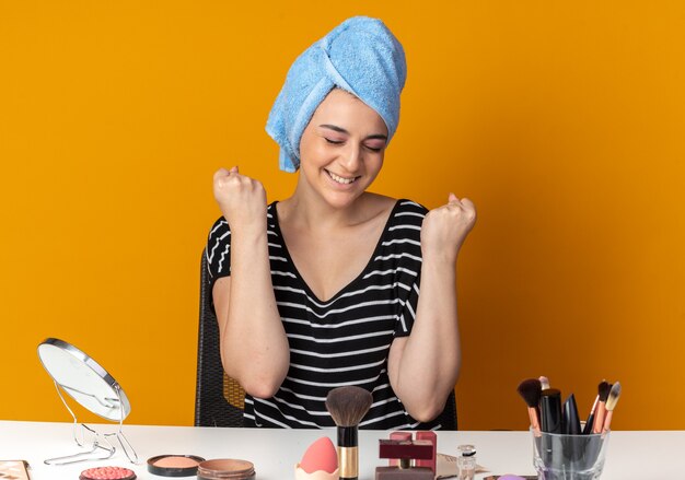 Foto gratuita soddisfatto degli occhi chiusi, la giovane bella ragazza si siede al tavolo con gli strumenti per il trucco avvolti i capelli in un asciugamano mostrando sì gesto isolato sul muro arancione orange