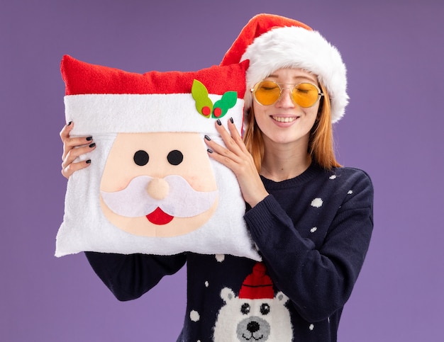 目を閉じて満足している若い美しい少女は、紫色の背景で隔離のクリスマス枕を保持しているメガネとクリスマスセーターと帽子を身に着けています