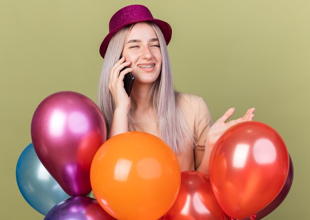 Pleased with closed eyes young beautiful girl wearing dental braces with party hat standing behind balloons speaks on phone 