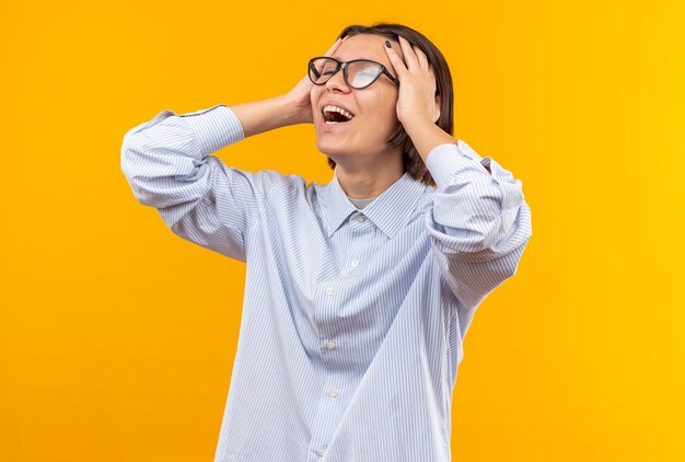 Pleased with closed eyes young beautiful girl wearing glasses grabbed head isolated on orange wall