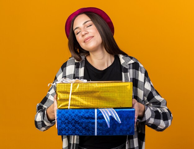 Pleased with closed eyes young beautiful woman wearing party hat holding gift boxes isolated on orange wall