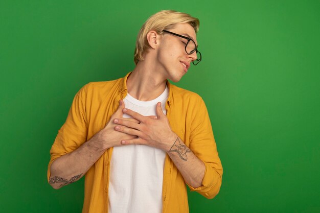 Pleased with closed eyes young blonde guy wearing yellow t-shirt and glasses putting hands on heart isolated on green with copy space