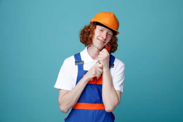 Pleased with closed eyes young builder man in uniform holding gas wrench isolated on blue background