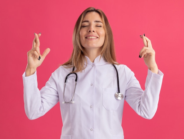 Довольная закрытыми глазами молодая женщина-врач в медицинском халате со стетоскопом, скрещивающим пальцы, изолирована на розовой стене