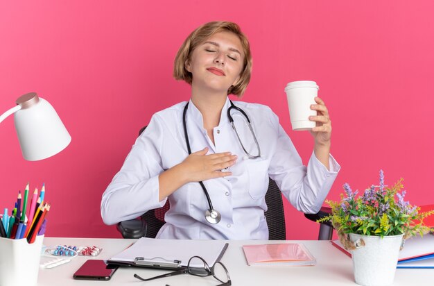 Pleased with closed eyes young female doctor wearing medical robe with stethoscope and glasses sits at desk with medical tools holding cup of coffee isolated on pink wall