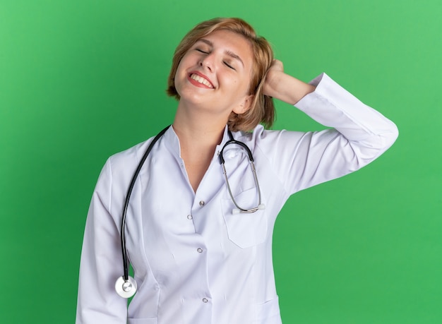 緑の壁に隔離された頭に手を置く聴診器で医療ローブを身に着けている目を閉じて満足している若い女性医師