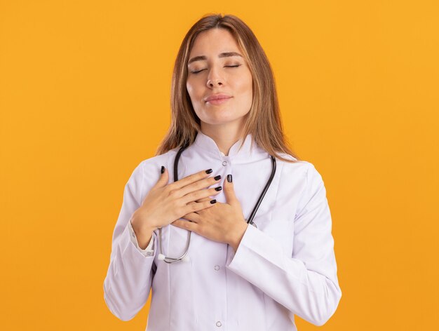 Pleased with closed eyes young female doctor wearing medical robe with stethoscope putting hands on heart isolated on yellow wall