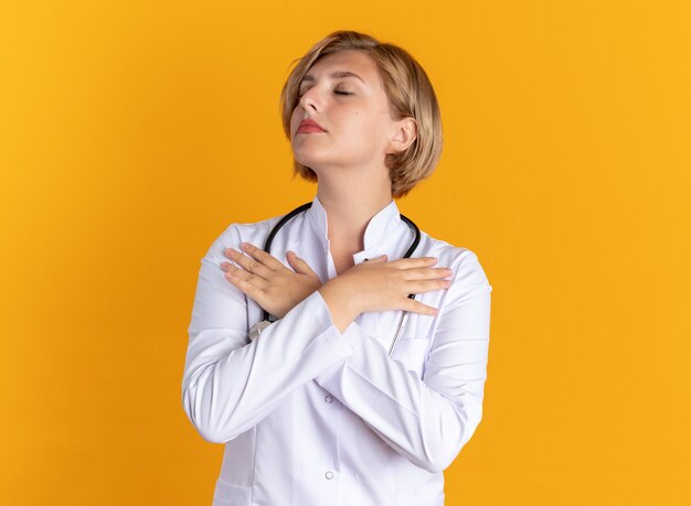 Pleased with closed eyes young female doctor wearing medical robe with stethoscope putting hands on shoulder isolated on orange wall
