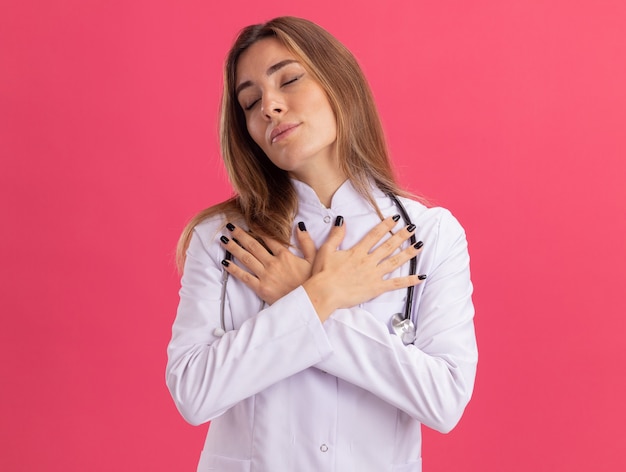 Pleased with closed eyes young female doctor wearing medical robe with stethoscope putting hands on shoulder isolated on pink wall